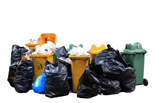 A collection of various rubbish bins and bags positioned outdoors on pavement, featuring a yellow wheelie bin with an open lid, filled with white and black waste, and a green wheelie bin to its right, both placed against a white background. Several large black waste bags are stacked in front and around the bins, some appearing partially open or tied. There is also a blue bag among the waste, and the bins are made of plastic with textured finishes. The scene appears to be part of a rubbish removal process related to office clearance or general waste disposal in Erith or nearby areas, as indicated by the context of the website. The lighting is consistent with natural daylight, highlighting the different textures and colors of the waste containers and bags. This setup reflects typical arrangements for waste collection and removal services, such as those offered by Office Clearance Erith, with a focus on clearing debris from commercial or residential settings in the local postcode area.