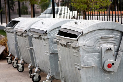 Four large, industrial-style metal wheelie bins are lined up on a paved outdoor surface, with their lids closed. The bins are constructed from galvanized steel, exhibiting a textured, slightly reflective metallic finish with visible ridges and circular patterns on the sides. The bins are positioned in a row, with the nearest bin on the right slightly ahead of the others, which are aligned in parallel behind it. Each bin has four small caster wheels at the base, facilitating movement, and are fitted with lids that are hinged at the back, with the front edges visible. The environment appears to be a paved street or driveway, with a metal fence and parked cars visible in the background, along with some greenery from trees or shrubs. The setting suggests an urban or suburban area within or near Erith, where Office Clearance Erith offers rubbish removal services. The lighting indicates daytime with natural, diffuse illumination, highlighting the metallic finish and practical design of the waste bins.
