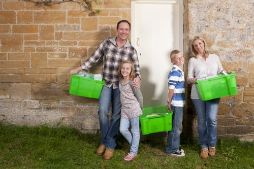 A group of four people, consisting of a man, woman, and two children, standing outdoors against a weathered brick wall. The man is holding a bright green plastic storage box in his right hand, while the woman on the right holds a similar box in her left hand. The girl in the center has a matching green box placed on the ground in front of her, and the boy is standing beside her, holding a green box. The family appears to be engaged in a rubbish removal activity, with all members smiling. They are on a patch of grass near the wall, which has patches of stone and brick with visible texture and weathering, suggesting an external setting, possibly near a residential or commercial property in Erith. The scene is evenly lit with natural light, highlighting the vivid green of the storage boxes and the casual clothing of the family, supporting the context of rubbish collection or clearance services by Office Clearance Erith.