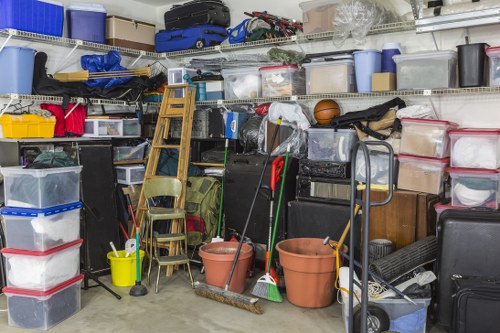 A densely packed storage area or garage space filled with various plastic storage containers, some transparent and others opaque, stacked on metal shelving units and on the floor. The containers are in different sizes and colors, including clear, blue, red, and black, with some lids visible, indicating they are used for storing household or office items. In the foreground, there are two large terracotta flower pots, a small wooden ladder leaning against the shelving, and several cleaning tools such as mops and brooms placed upright. A few folding chairs are positioned next to the containers, and a black trolley or hand truck is visible on the right side. The environment appears utilitarian, with a concrete floor and artificial lighting, typical of a garage or storage room used for holding clutter or items awaiting disposal. This image aligns with the services offered by Office Clearance Erith, who specialise in rubbish removal efforts in the local area around Erith, Kent.