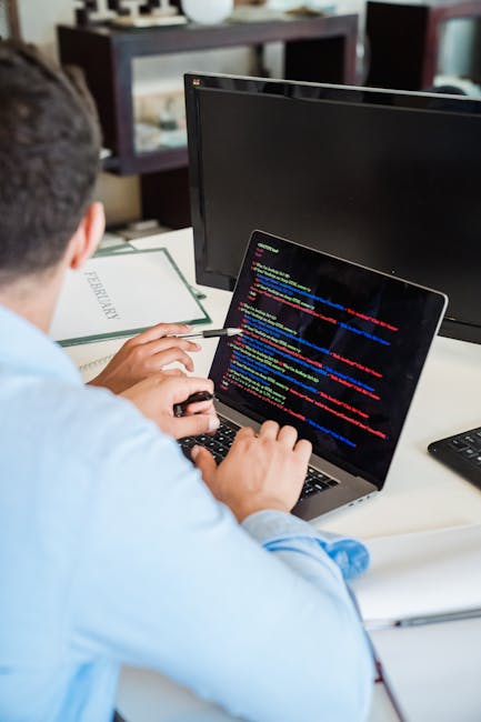 A person working at a desk with a laptop displaying lines of colorful computer code. The individual is seated with their back to the camera, wearing a light blue shirt, and holding a mouse in their ri
