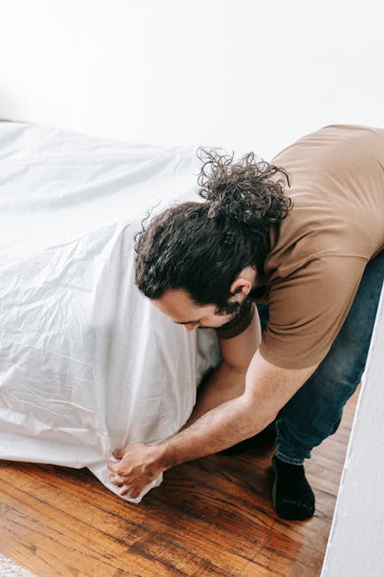A man with dark, curly hair, dressed in a brown T-shirt and dark jeans, is kneeling on a wooden floor beside a bed in a well-lit room with plain white walls. He is holding and adjusting a large white 